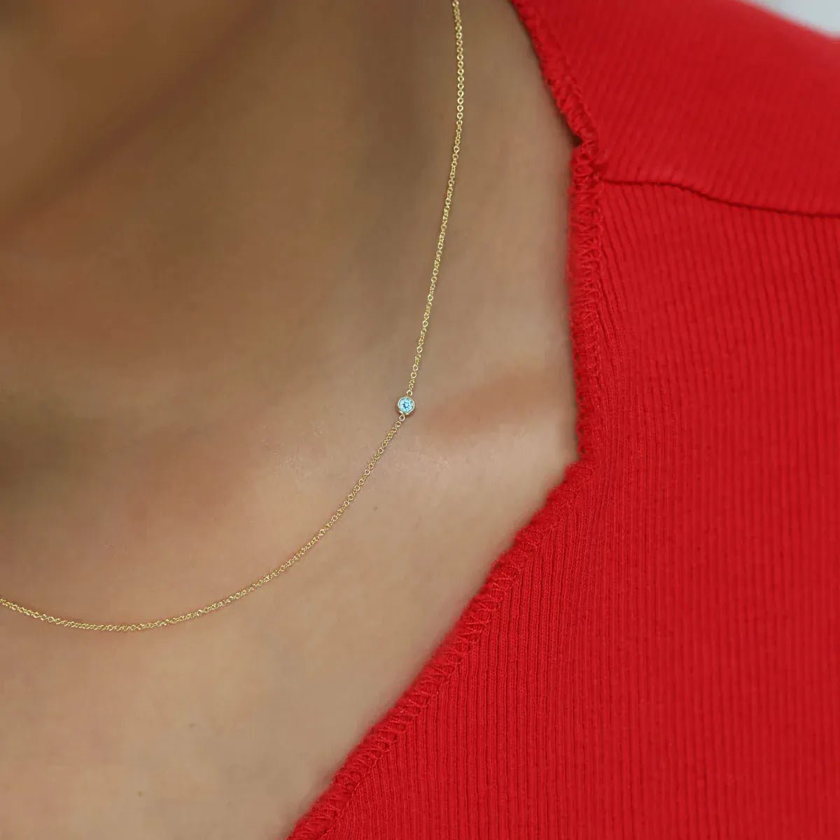 A close-up of a neck wearing a yellow gold 14K Gold Asymmetrical Birthstone Necklace - Aquamarine (March), styled with a red top. 