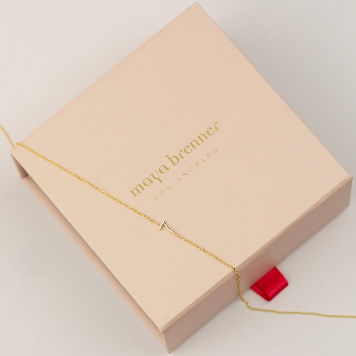 A 14k Gold Asymmetrical Number Necklace - 7 in yellow gold, laid on top of a light pink Maya Brenner jewellery box, on a white background. 