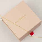 A 14k Gold Asymmetrical Number Necklace - 7 in yellow gold, laid on top of a light pink Maya Brenner jewellery box, on a white background. 