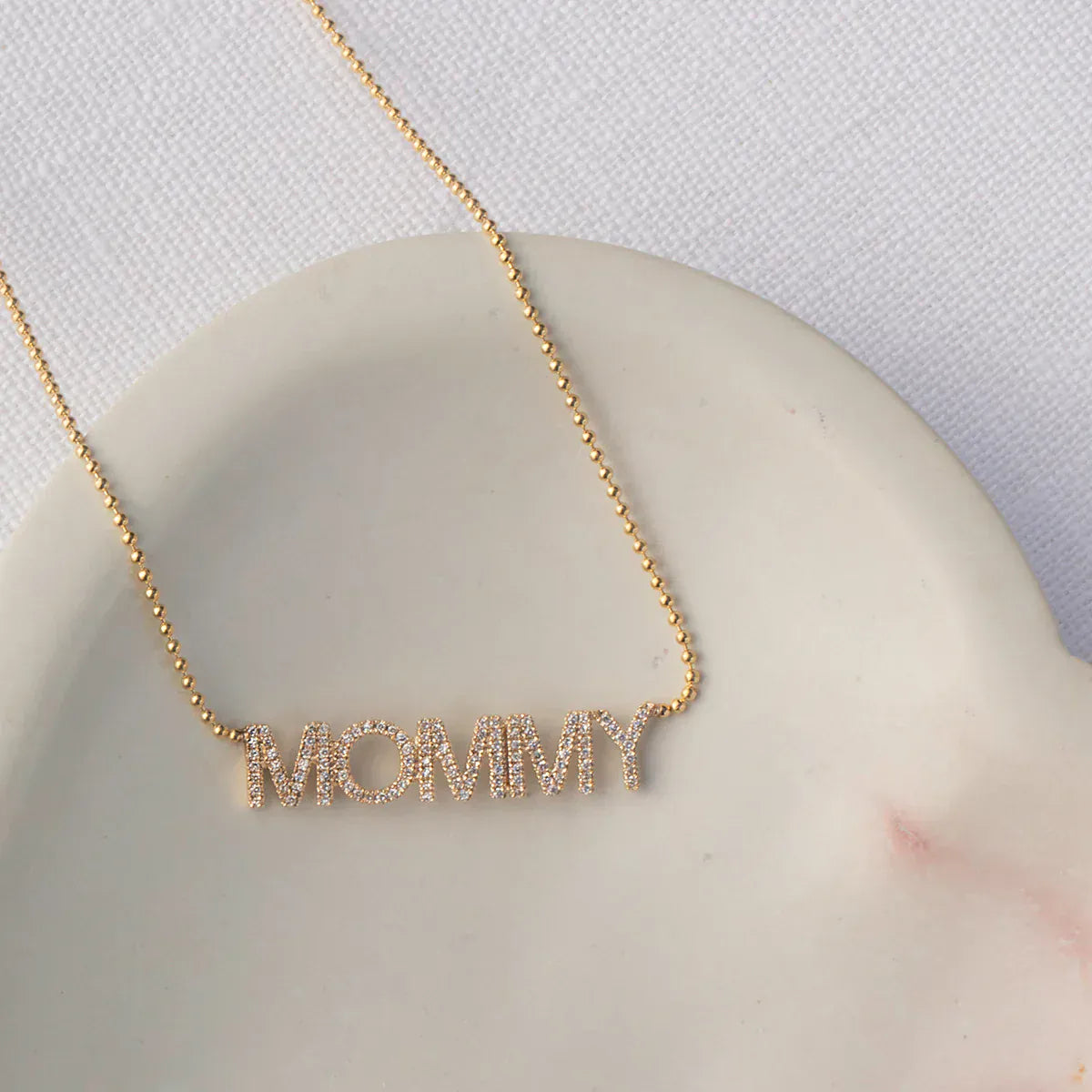 A Pavé MOMMY Necklace in yellow gold on a cable chain laid on top of a cream surface and white fabric background. 