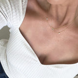Close-up of a person wearing an off-white top and three yellow gold necklaces – one with a letter V charm, one with a letter O charm, and one with a birthstone.