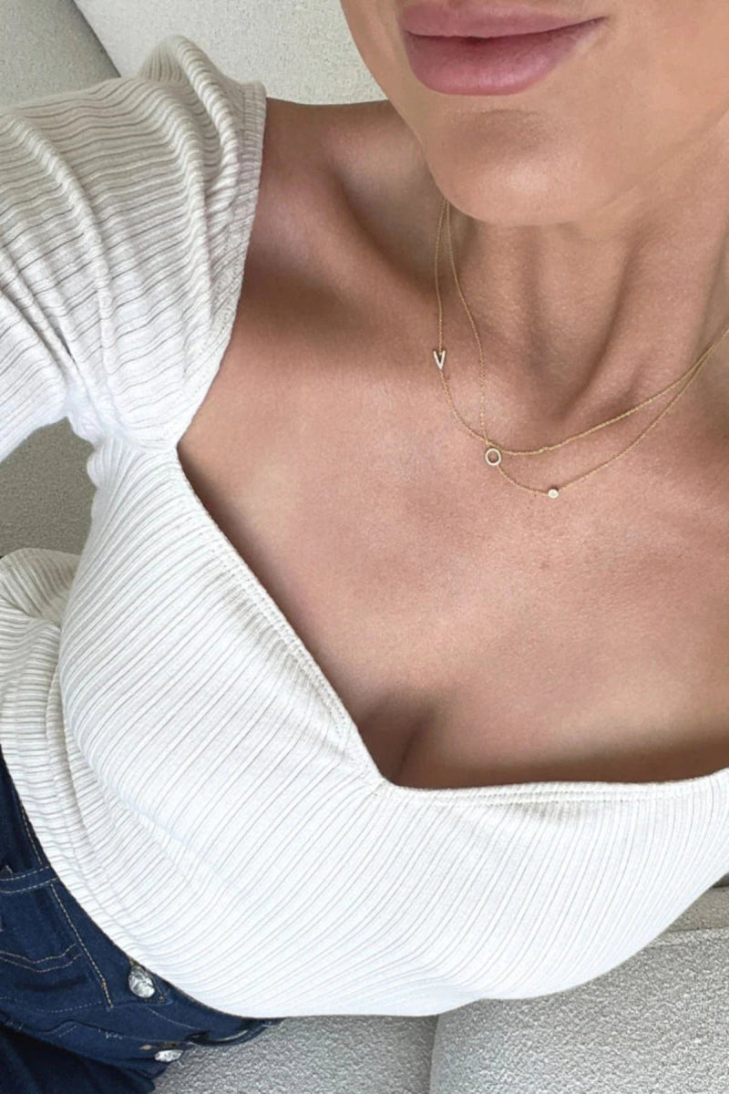 Close-up of a person wearing an off-white top and three yellow gold necklaces – one with a letter V charm, one with a letter O charm, and one with a birthstone.