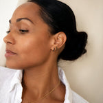 A woman wearing two Alli Webb Onyx Huggies and gold necklaces with a white shirt against a light background. 