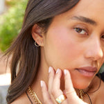 Close-up of a woman wearing a yellow gold Open Eclipse Earring - Small, Thick Band and several necklaces, with brown hair and an outdoors blurred background. 