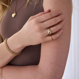 Close-up of a hand wearing a yellow gold Paris Ring, a Tube Ring and two Rolo Bracelets, styled with a brown top and the Peony Bouquet Necklace with a Diamond Heart Locket Charm, against a white background.