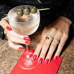Close-up of a pair of hands wearing a Make it a Double Ring, Cosmo Ring, Open Diamond and two Pave diamond Rings with red nails holding a cocktail. 