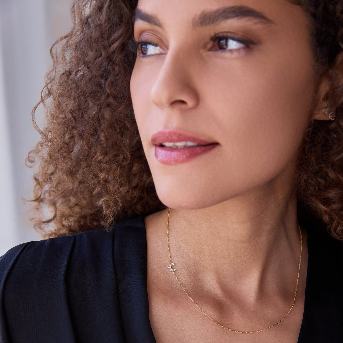 Close-up of a woman wearing a yellow gold Custom Gold Necklace - 1 Letter with a pave moon charm, with a navy blue top and brown curly hair, against a blurred background. 