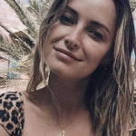 A blonde woman wearing an England pendant necklace in yellow gold and a  leopard print top with palm trees in the background.