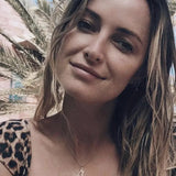 A blonde woman wearing an England pendant necklace in yellow gold and a  leopard print top with palm trees in the background.