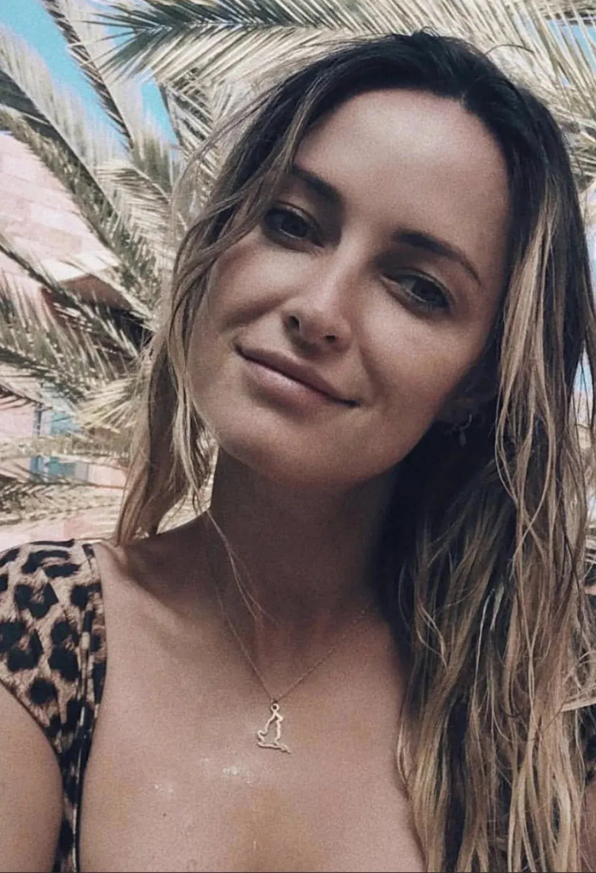 A blonde woman wearing an England pendant necklace in yellow gold and a  leopard print top with palm trees in the background.