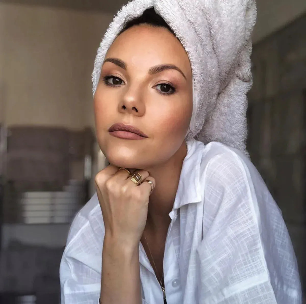 A woman wearing several rings, including a Perla Pinky Ring with a towel  wrapped around her head, wearing a white shirt in a blurred indoor setting. 