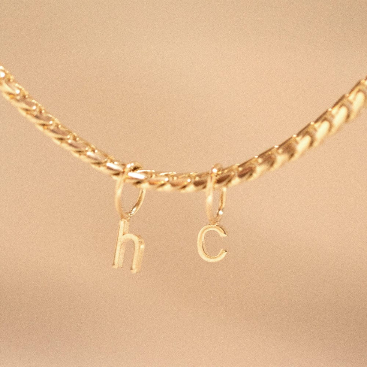 A yellow gold necklace with Petite Letter Charms in h and c on against a beige background.