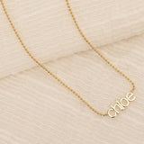 A yellow gold Petite Nameplate Necklace with 'chloe' against a cream fabric background. 