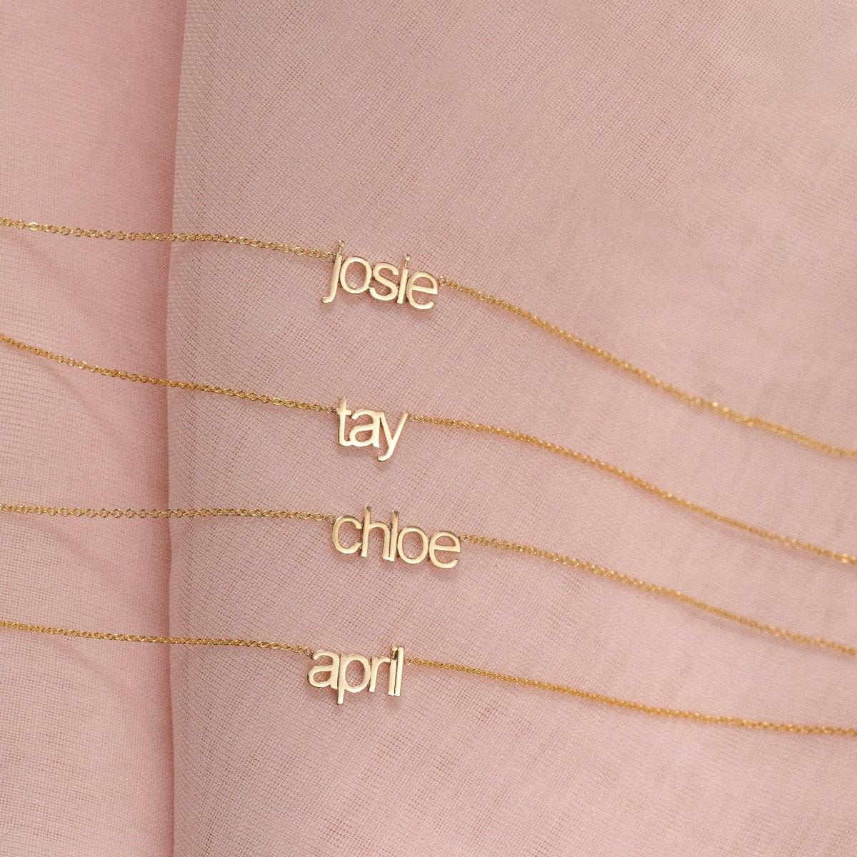 Four Petite Nameplate Necklaces in yellow gold, in the names Josie, Tay, Chloe and April (all in lowercase) on a pink fabric background. 