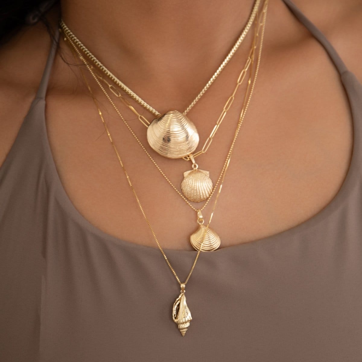 A close-up of a woman wearing a Chic Cuban Curb Chain with The Golden Venus Shell, an Element Long Link Chain with a Golden Scallop Shell, a Classic Bead Chain with a Golden Coast Shell, and The Slide Chain with a Golden Spiral Shell, with a light brown singlet top. 