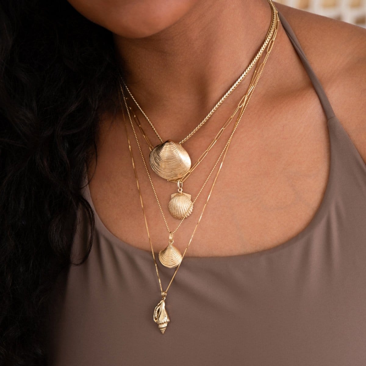 A close-up of a woman wearing a Chic Cuban Curb Chain with The Golden Venus Shell, an Element Long Link Chain with a Golden Scallop Shell, a Classic Bead Chain with a Golden Coast Shell, and The Slide Chain with a Golden Spiral Shell, with a light brown singlet top. 
