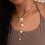 A close-up of a woman wearing a Chic Cuban Curb Chain with The Golden Venus Shell, an Element Long Link Chain with a Golden Scallop Shell, a Classic Bead Chain with a Golden Coast Shell, and The Slide Chain with a Golden Spiral Shell, with a light brown singlet top. 