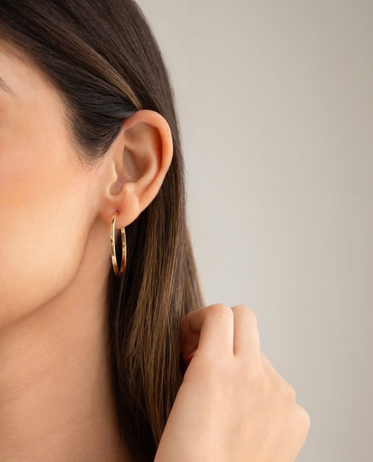 Close-up of woman's ear wearing Sleek & Chic Hoops - Small in yellow gold with her brown hair visible against a light grey background. 