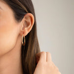 Close-up of woman's ear wearing Sleek & Chic Hoops - Small in yellow gold with her brown hair visible against a light grey background. 