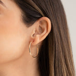 Close-up of woman's ear wearing Sleek & Chic Hoops - Small in yellow gold with her brown hair visible against a light grey background. 
