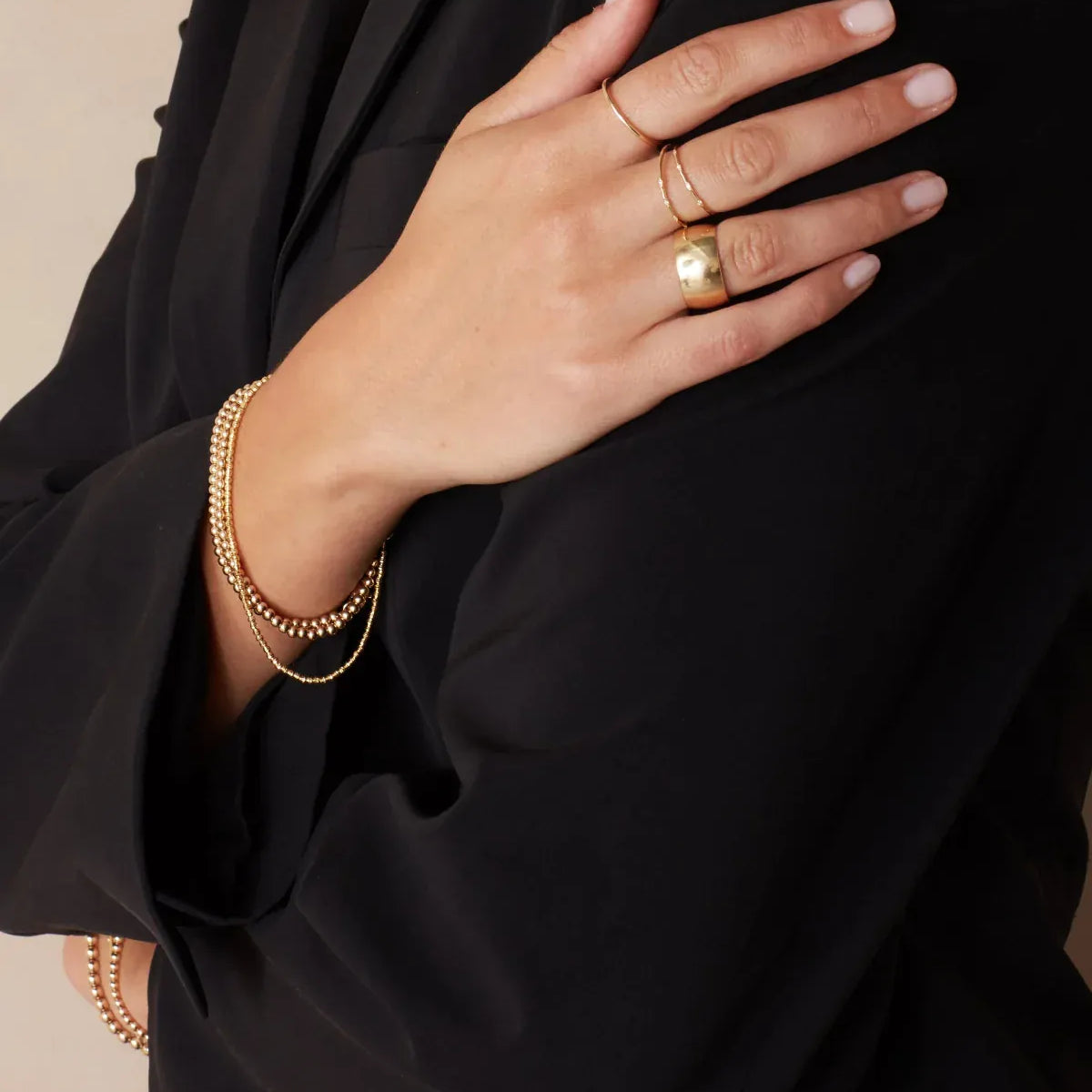 A woman's hand wearing two 4 Diamond Rings, a Thick Band and Delicate Band, with Bubble Bracelets and a Diamond Cut Bracelet, all in yellow gold, resting on her black blazer against a light grey background. 