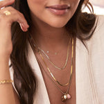 Close-up of a woman wearing multiple yellow gold necklaces, including a Custom Gold Necklace with the letters K and R, an Element Short Link Chain, an Overboard Layering Chain, and a Round Curb Chain with a Pink Opal Tear Drop Charm, an Engraved Heart Charm, and a Diamond Heart Locket Charm, paired with a cream top.