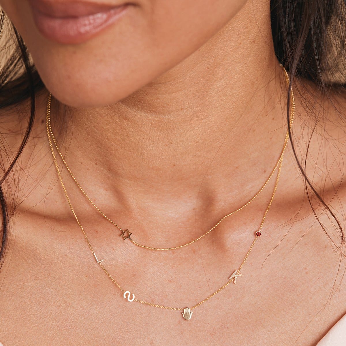 A close-up of a woman’s neck wearing a Custom Gold Necklace with five charms (letters L, S, and K, a hand and a birthstone) layered with a Charm Necklace featuring a Star of David charm, both in yellow gold.