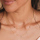 A close-up of a woman’s neck wearing a Custom Gold Necklace with five charms (letters L, S, and K, a hand and a birthstone) layered with a Charm Necklace featuring a Star of David charm, both in yellow gold.