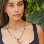 A woman wearing multiple necklaces, including a Custom Gold Necklace with four letters, an Element Short Link Chain, a Rolo Necklace, a Black Rhodium Antique Beach Chain and a Round Curb Chain with a Diamond Heart Locket, an Etched Turquoise Heart Charm and a Balance Crystal Charm, along with Open Eclipse Earrings and a black top.