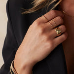 A woman’s hand wearing two 4 Diamond Rings, a Thick Band, a Delicate Band, Bubble Bracelets, a Diamond Cut Bracelet, and an Element Long Link Chain, all in yellow gold, styled with a black blazer against a light grey background.