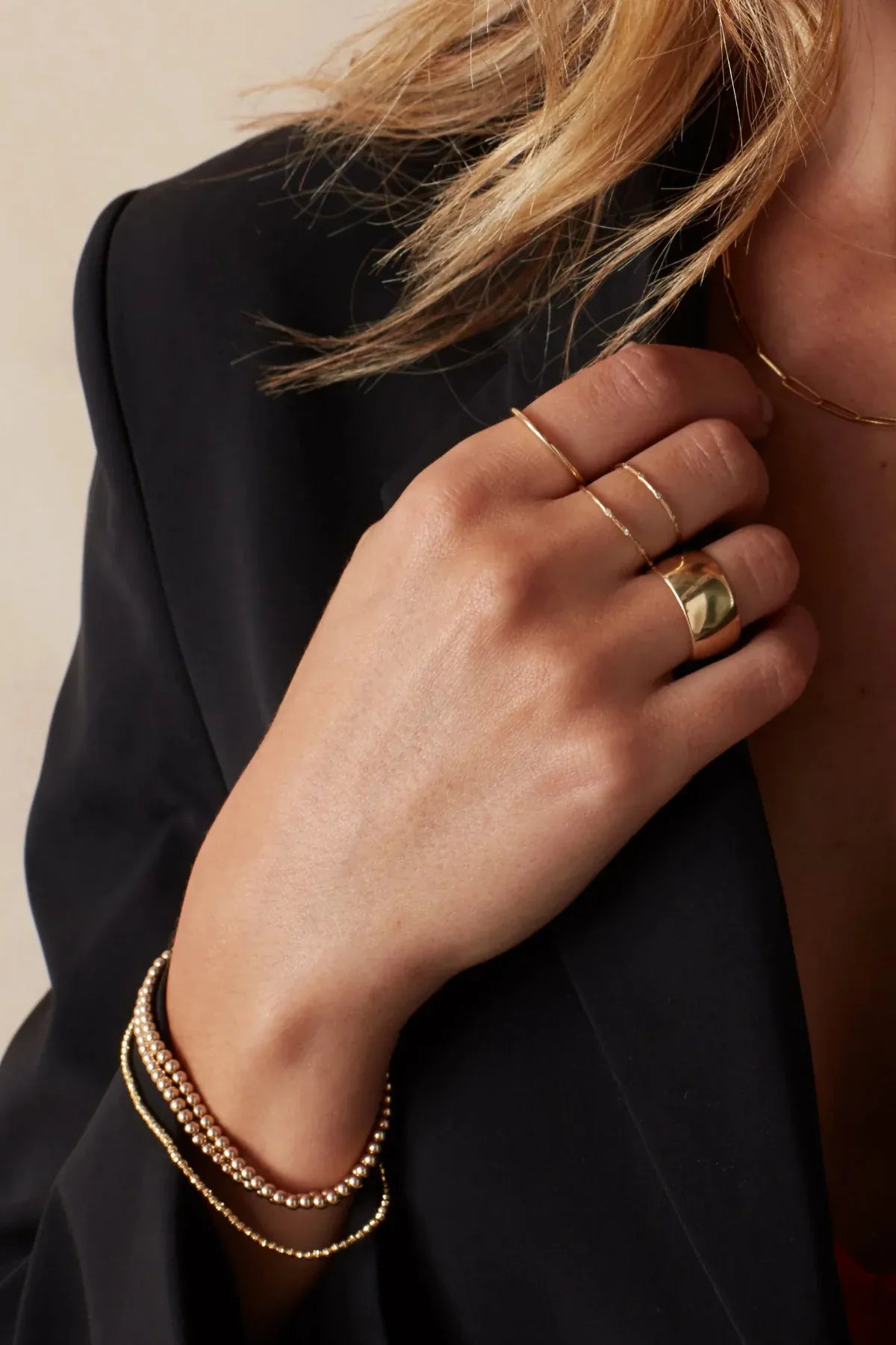 A woman’s hand wearing two 4 Diamond Rings, a Thick Band, a Delicate Band, Bubble Bracelets, a Diamond Cut Bracelet, and an Element Long Link Chain, all in yellow gold, styled with a black blazer against a light grey background.