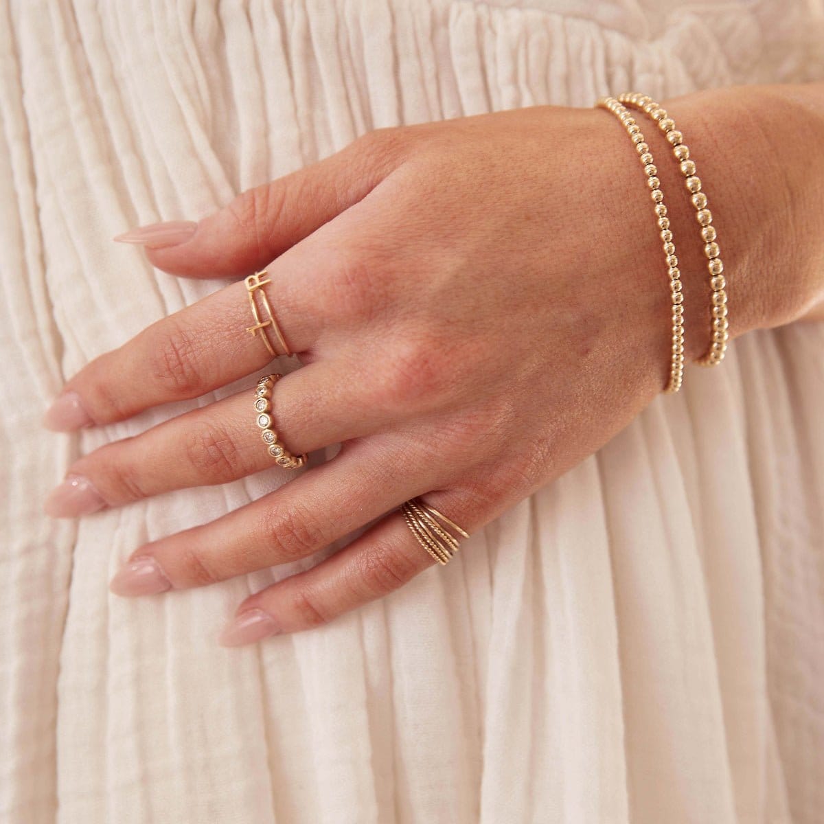 A woman’s hand resting on her cream dress, wearing gold jewellery including Bubble Bracelets, an R Letter Ring, a Number 1 Ring, Delicate Trio Stacking Rings, and a Magnolia Bouquet Ring.