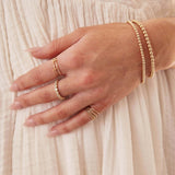 A woman’s hand resting on her cream dress, wearing gold jewellery including Bubble Bracelets, an R Letter Ring, a Number 1 Ring, Delicate Trio Stacking Rings, and a Magnolia Bouquet Ring.