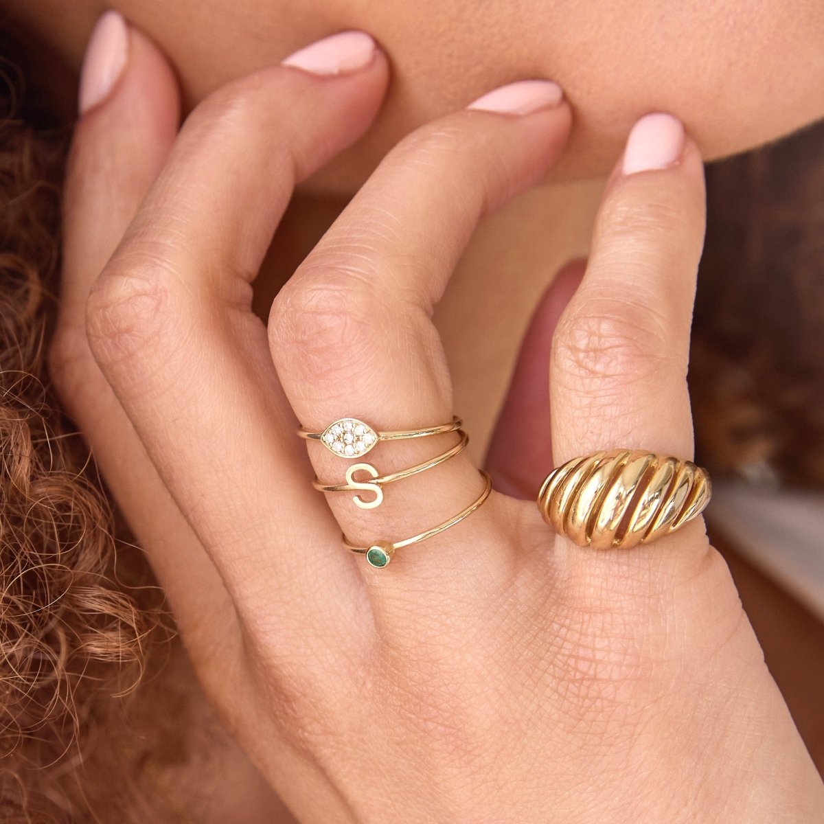 A close-up of a woman's hand wearing various yellow gold rings, including an Etched Paris Ring, a Pave Petal Ring, an S Letter Ring and a Birthstone Ring. 