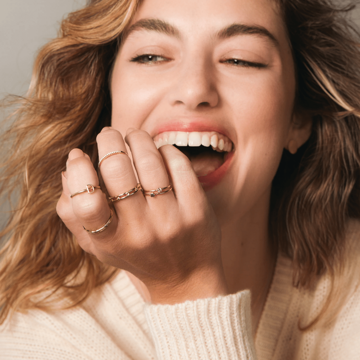 A woman with a joyful expression wearing multiple gold rings on her fingers, including a D Letter Ring, two Birthstone Rings, a Link Ring, a Delicate Band and a Pavé Diamond Ring.