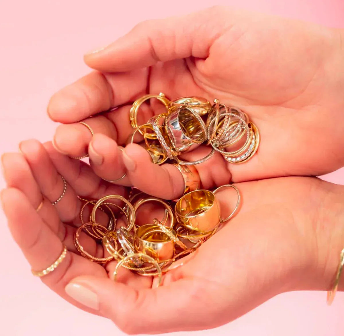 A pair of hands holding several yellow gold and white gold rings, including Thick Bands, Delicate Trio Stacking Rings, Braided Rings, Flat Rings, against a light pink background. 