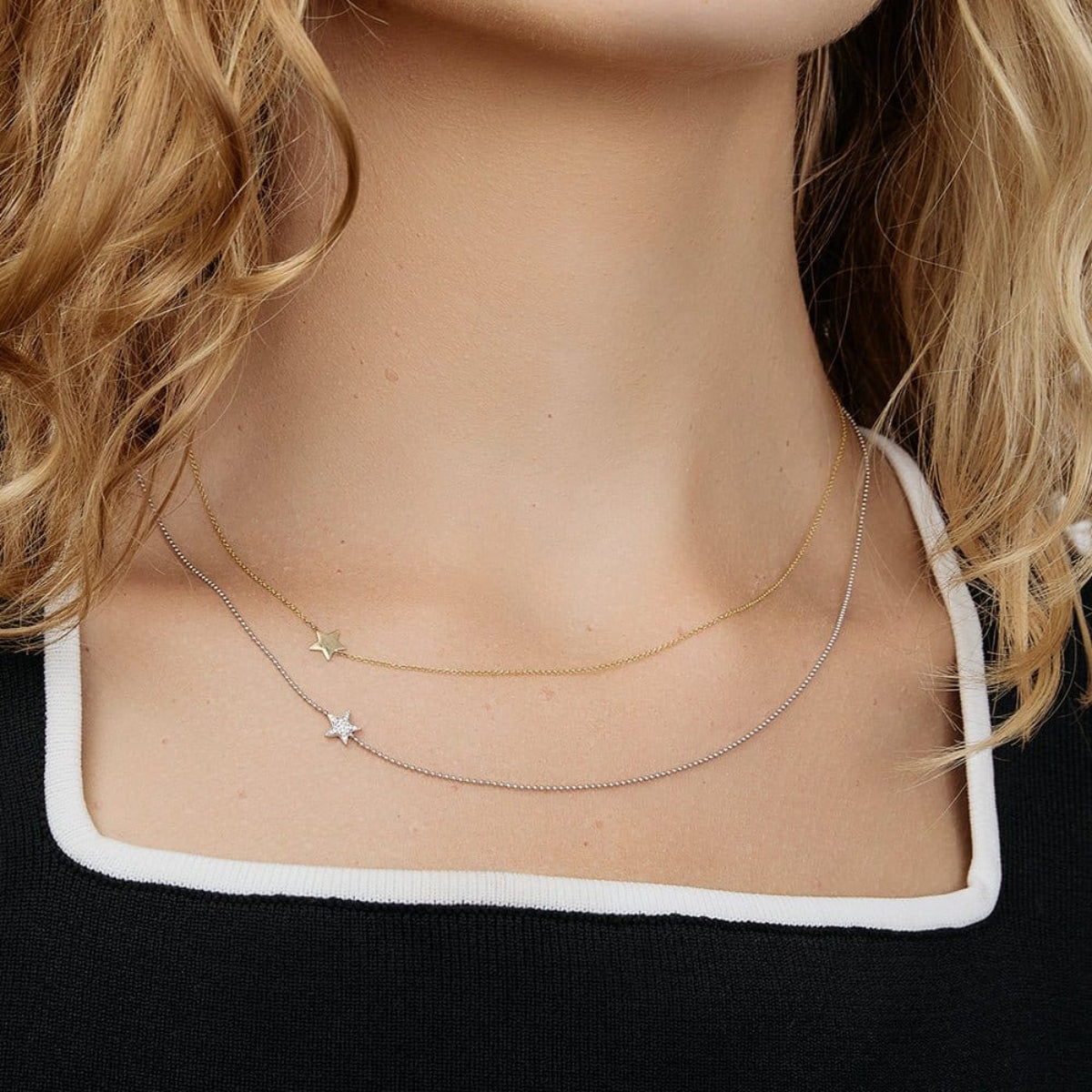 A close-up of a woman's neck wearing two 14k Gold Asymmetrical Charm Necklaces (one star in yellow gold and one pavé star in white gold), styled with a black and white top.