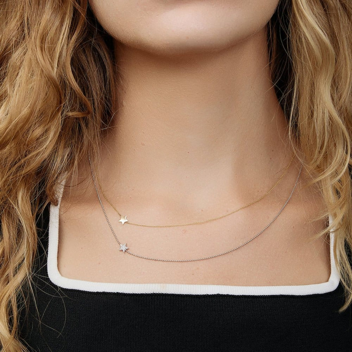 A close-up of a woman's neck wearing two 14k Gold Asymmetrical Charm Necklaces (one star in yellow gold and one pavé star in white gold), styled with a black and white top. 
