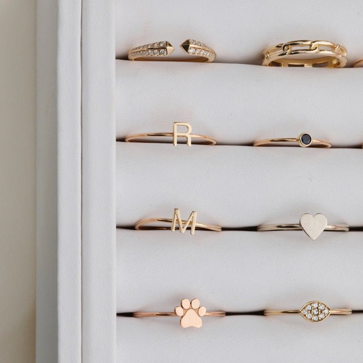 A number of yellow gold and rose gold rings in a white case, including an Open Diamond Ring, a Link Ring, an R Letter Ring, a Birthstone Ring, an M Letter Ring, a Heartfelt Ring, a paw ring, and a Pavé Petal Ring.
