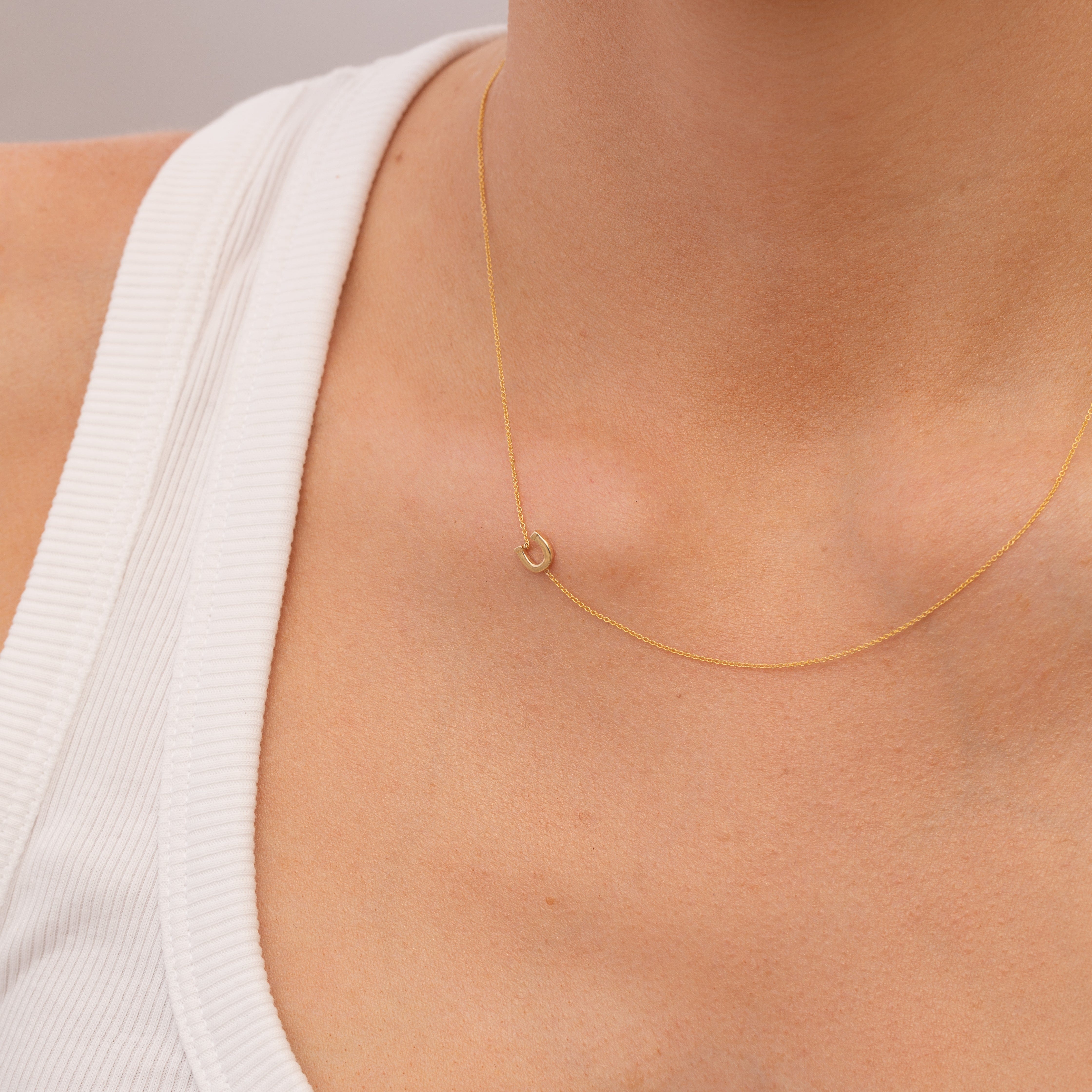 Gold necklace with a small horeshoe charm on a person wearing a white top against a neutral background