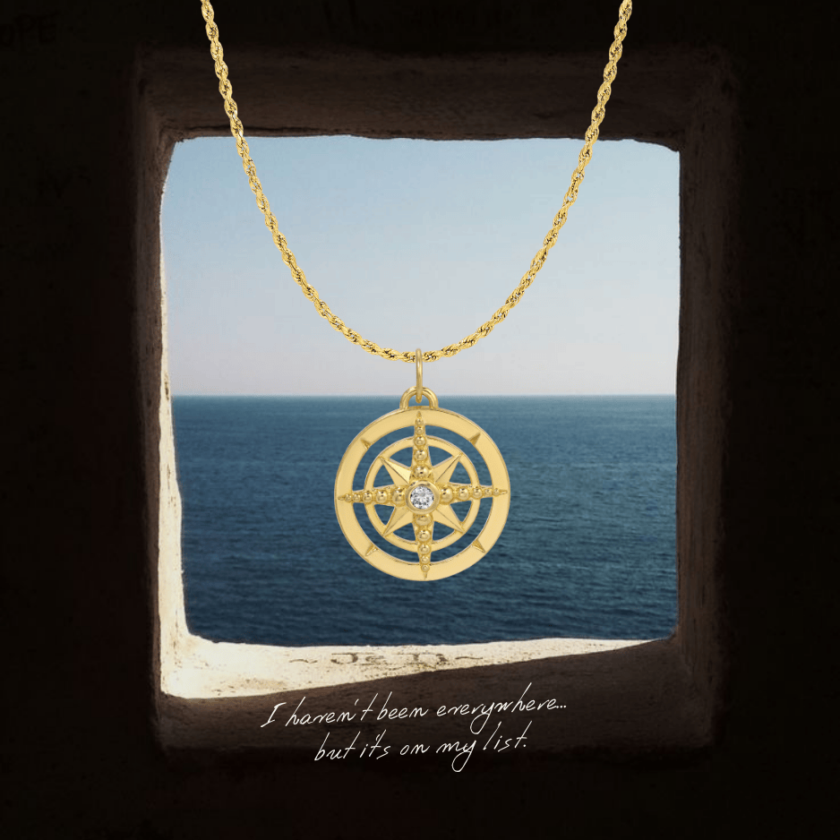 Gold compass necklace with a diamond on a stone ledge with ocean view