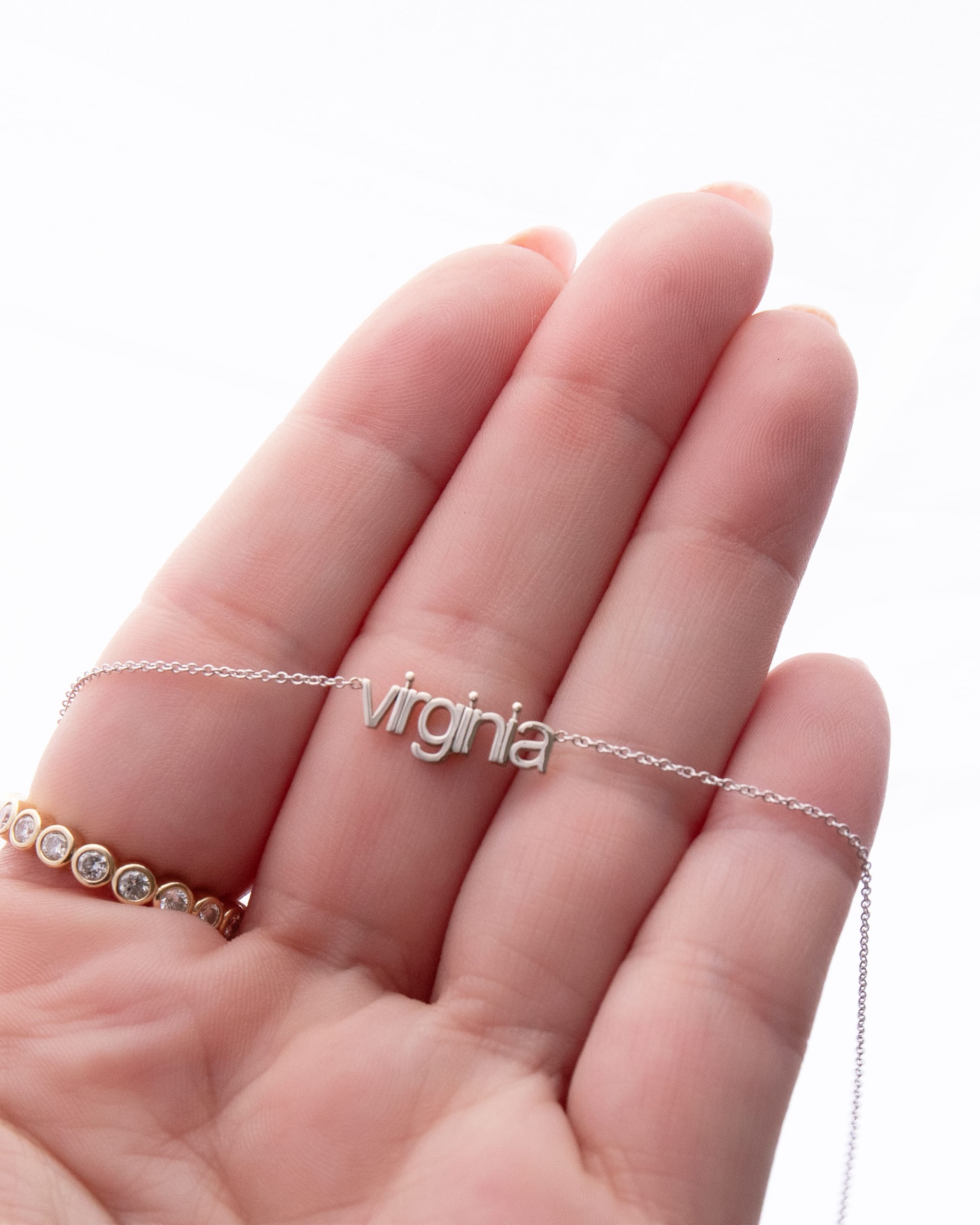Silver necklace with 'virginia' pendant on a hand against a white background