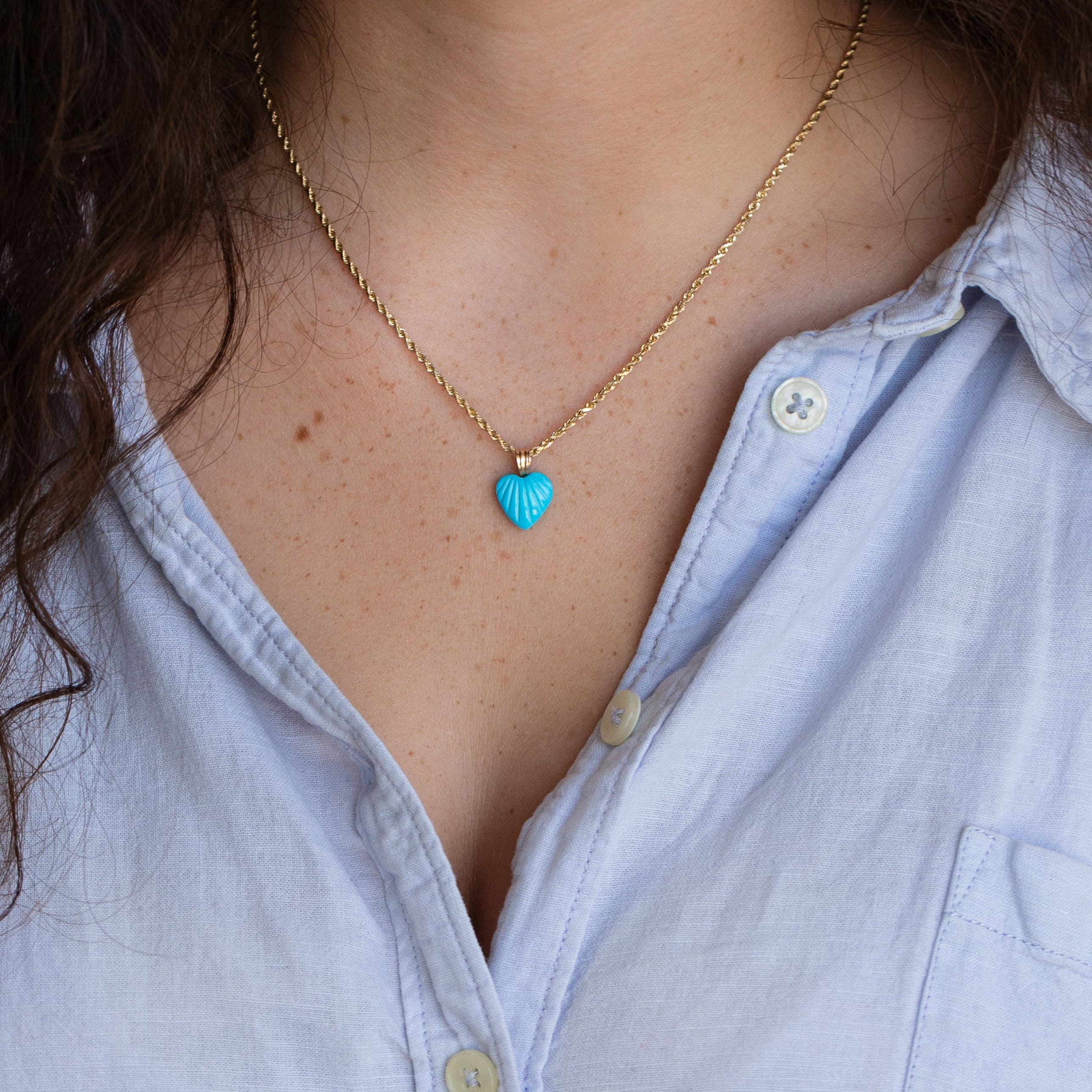 A close-up of a person wearing a delicate gold chain necklace with a small turquoise blue shell-shaped pendant, styled with a light blue button-down shirt.