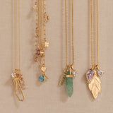 Four gold necklaces with unique charms are displayed on a beige fabric wall. The necklaces feature a dried flower, a blue gemstone, a green crystal, and a gold leaf pendant with a purple heart gemstone and clear stones.