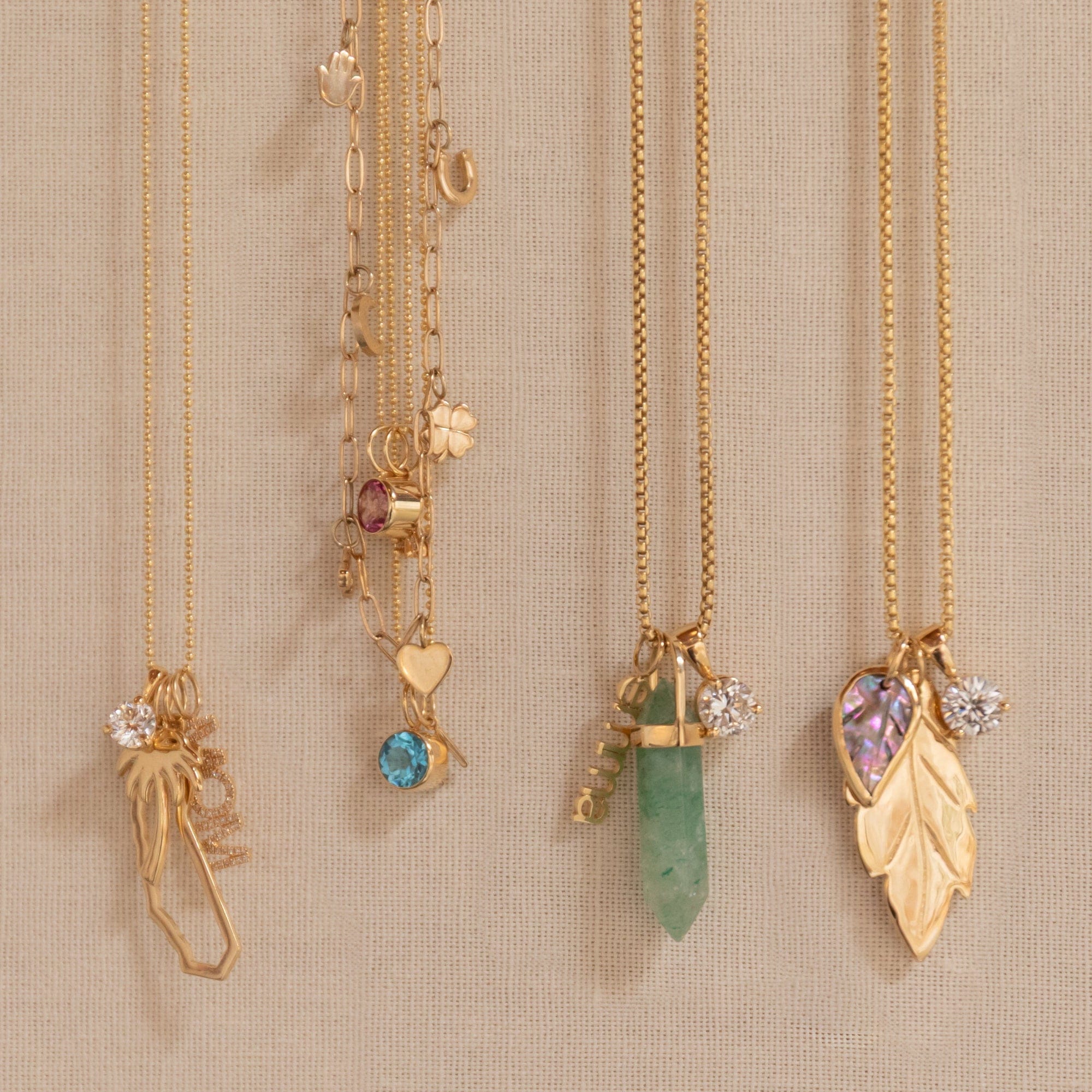 Four gold necklaces with unique charms are displayed on a beige fabric wall. The necklaces feature a dried flower, a blue gemstone, a green crystal, and a gold leaf pendant with a purple heart gemstone and clear stones.