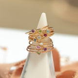 A stack of slim rose gold rings with small colorful gemstones, displayed on a white cone jewelry stand against a softly blurred warm beige background.