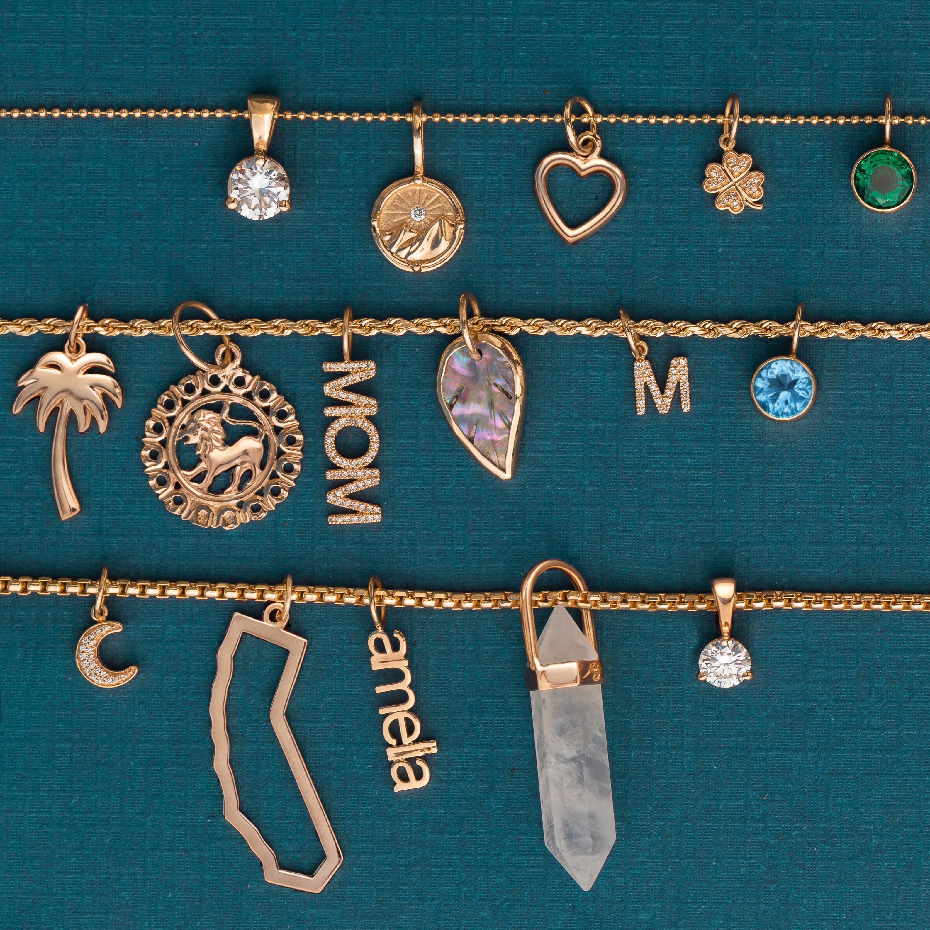 A collection of gold-tone jewelry charms, including a crescent moon, a palm tree, a lion emblem, the word 'MOM', a heart, a clover, a green gemstone, a blue gemstone, a crescent moon, a leaf, an 'M', a crystal point, and a nameplate reading 'amelia', displayed on thin chains against a textured teal background.