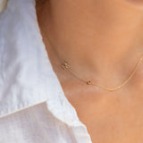 A delicate gold chain necklace worn on a model's neck, featuring a small cutout letter 'M' and a tiny round red gemstone accent, styled with a white textured blouse.