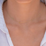 A close-up of a model's neck and chest wearing a delicate gold chain necklace with a small letter 'M' pendant and a tiny round yellow gemstone.
