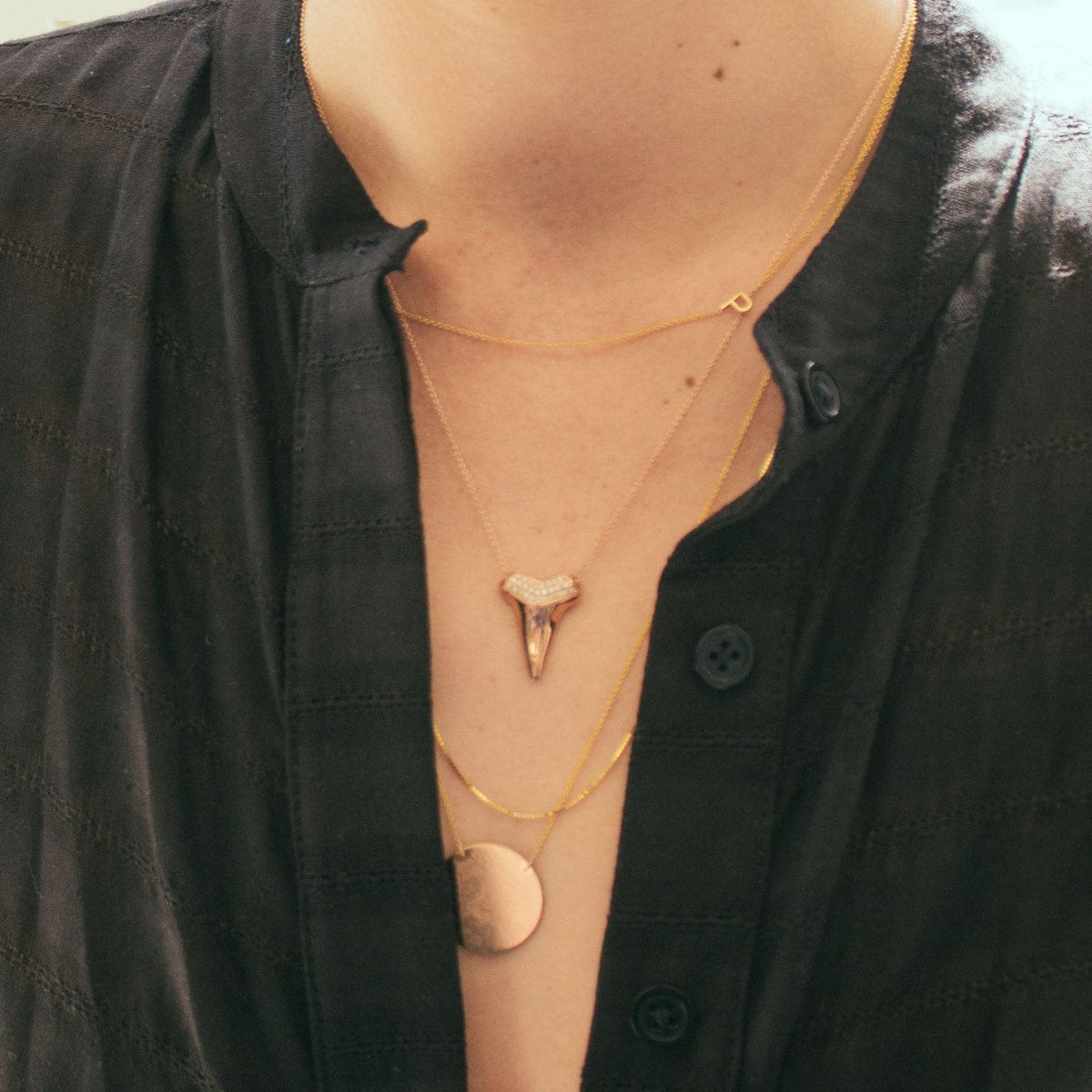 A close-up of a woman's neck wearing a black shirt and three rose gold necklaces: one with a letter P charm, one with a sharktooth with pavé on top, and one with a solid circle.